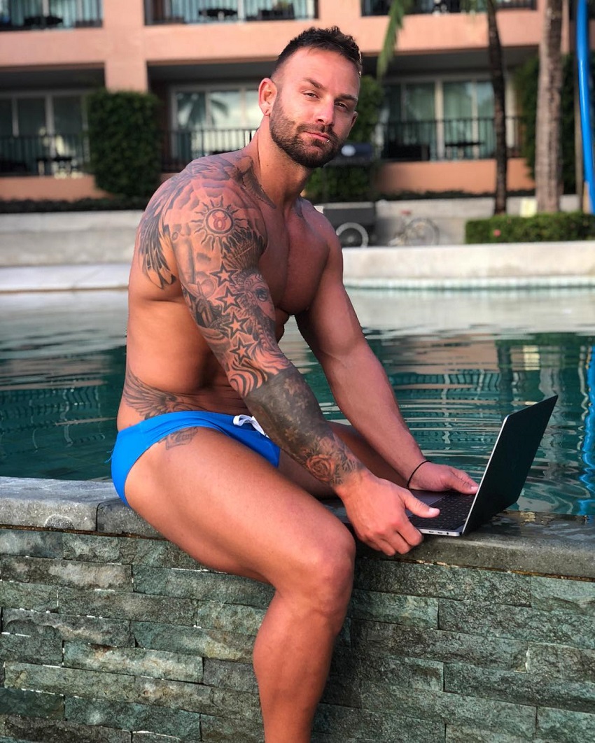 Joel Bushby sitting near a pool with his laptop, looking fit and muscular